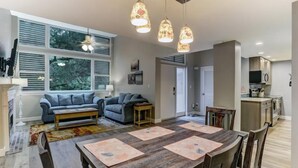 Un salon moderne avec un canapé, une table basse et une table à manger pour quatre personnes. Des luminaires suspendus sont placés au-dessus de la table, et une cheminée se trouve au fond de la pièce.