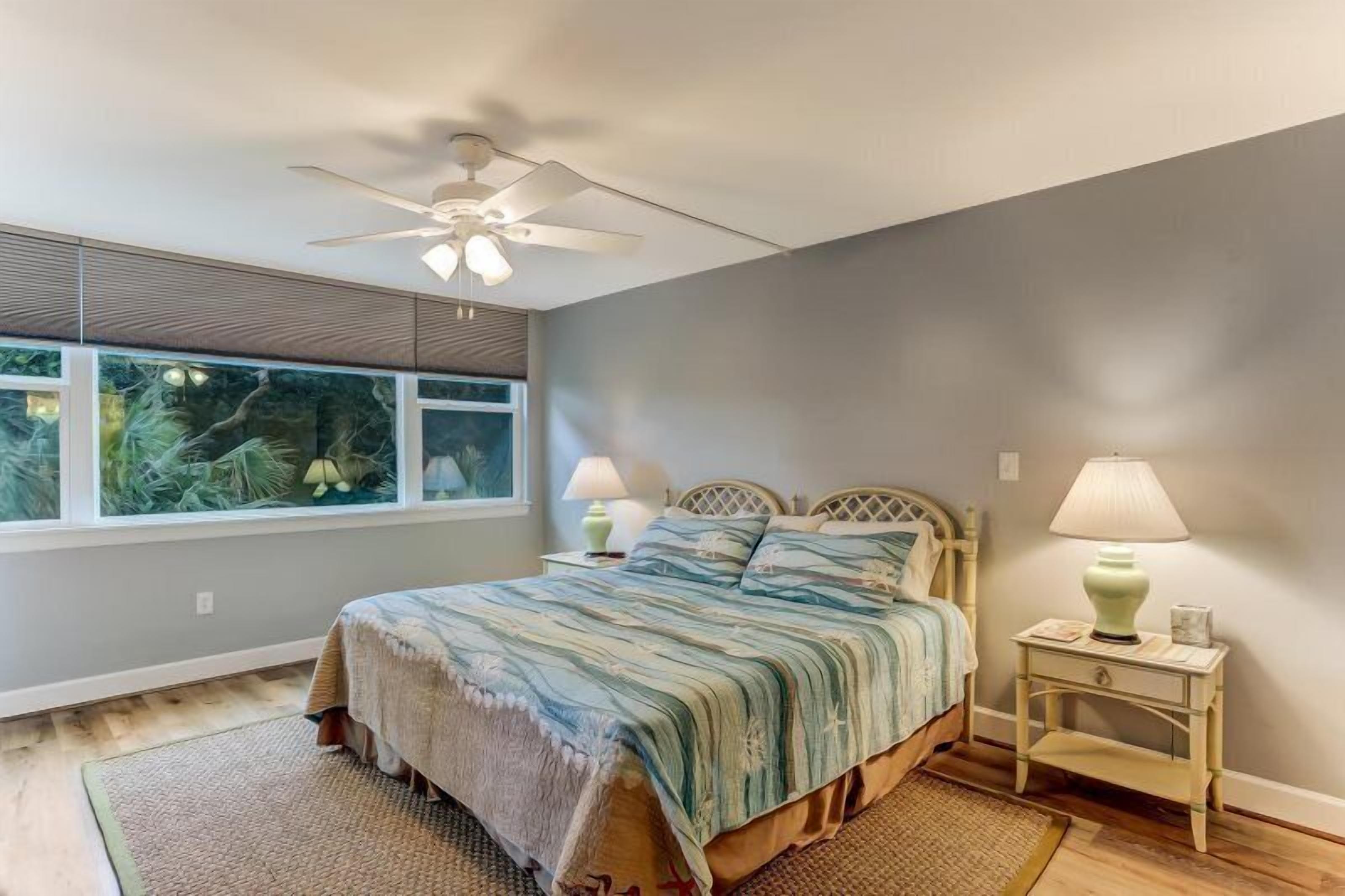 A bedroom with a bed, two bedside tables, a ceiling fan, and a window with a view of trees.