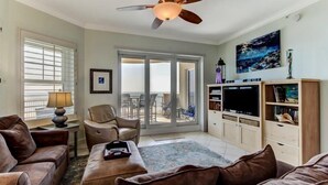 A living room with a ceiling fan, a sofa, a chair, a TV stand, and a view through the glass doors to the outside. - 61-ocean-place (Fernandina Beach)