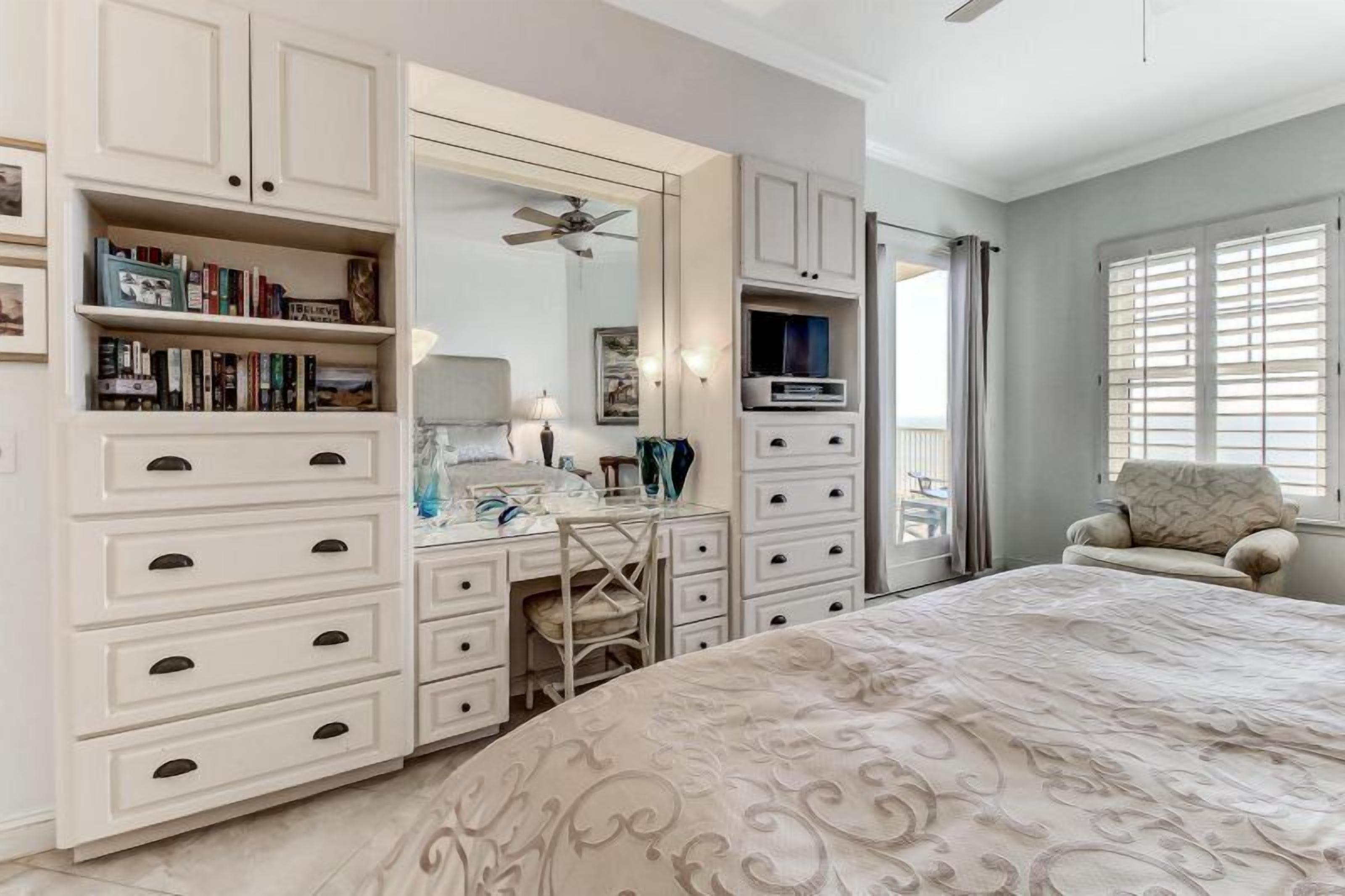 A bedroom with a bed, a desk, a chair, a wardrobe, a mirror, a ceiling fan, and a window with blinds.