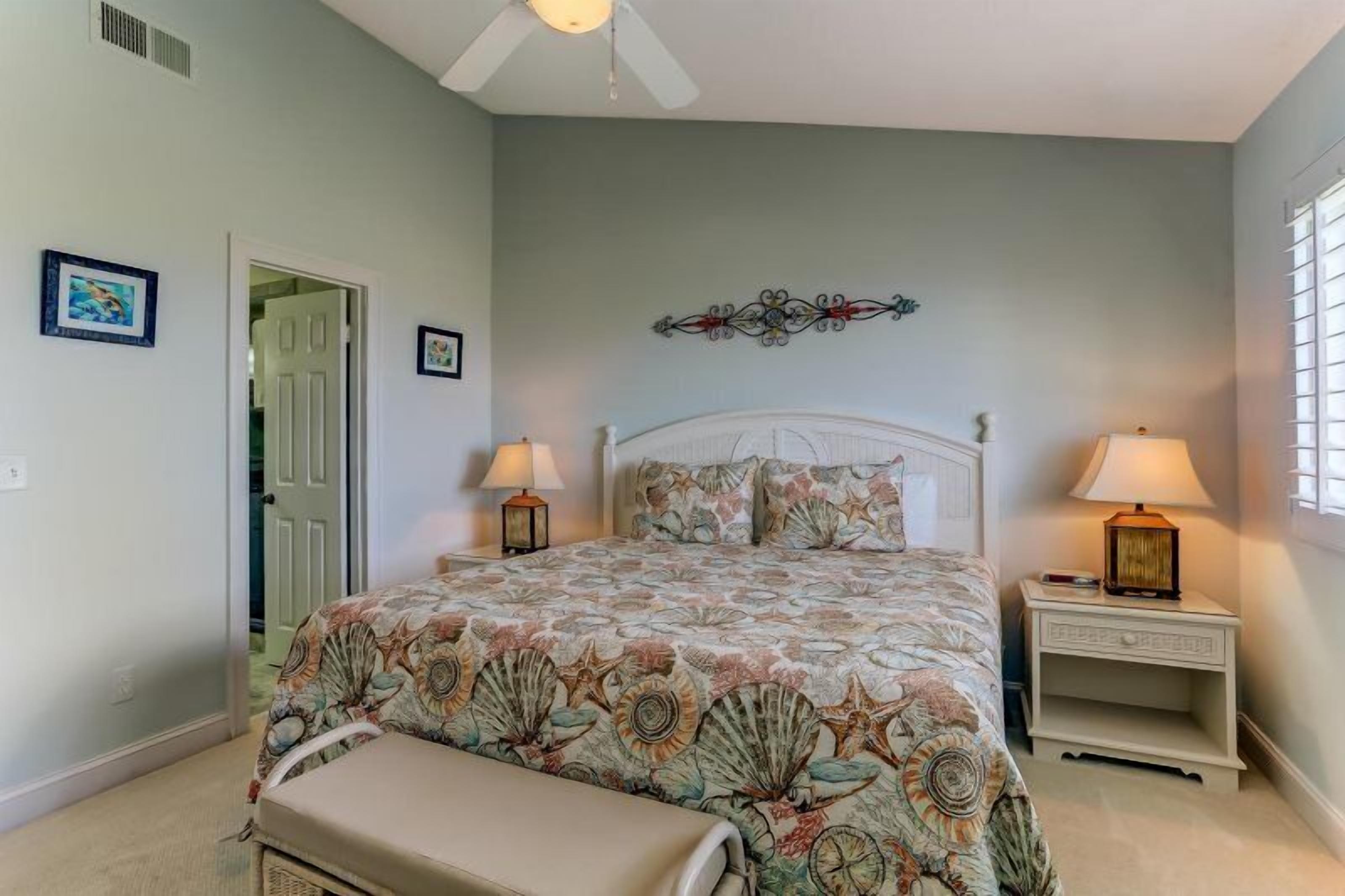 A bedroom with a bed, two bedside tables, a bench, a ceiling fan, and a door.