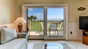 Una habitación con puerta de cristal corredera que da a un balcón con vista al océano y palmeras.
