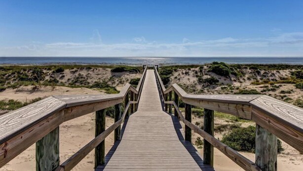 Un paseo marítimo de madera que conduce a una playa con dunas y un cielo despejado.