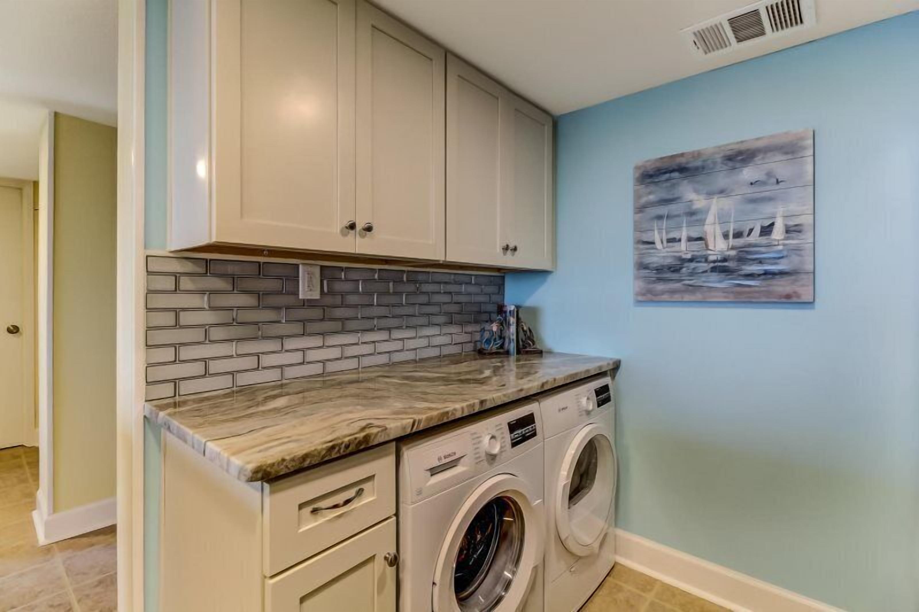 A laundry area with a washing machine, a tiled backsplash, and a painting of sailboats on the wall.