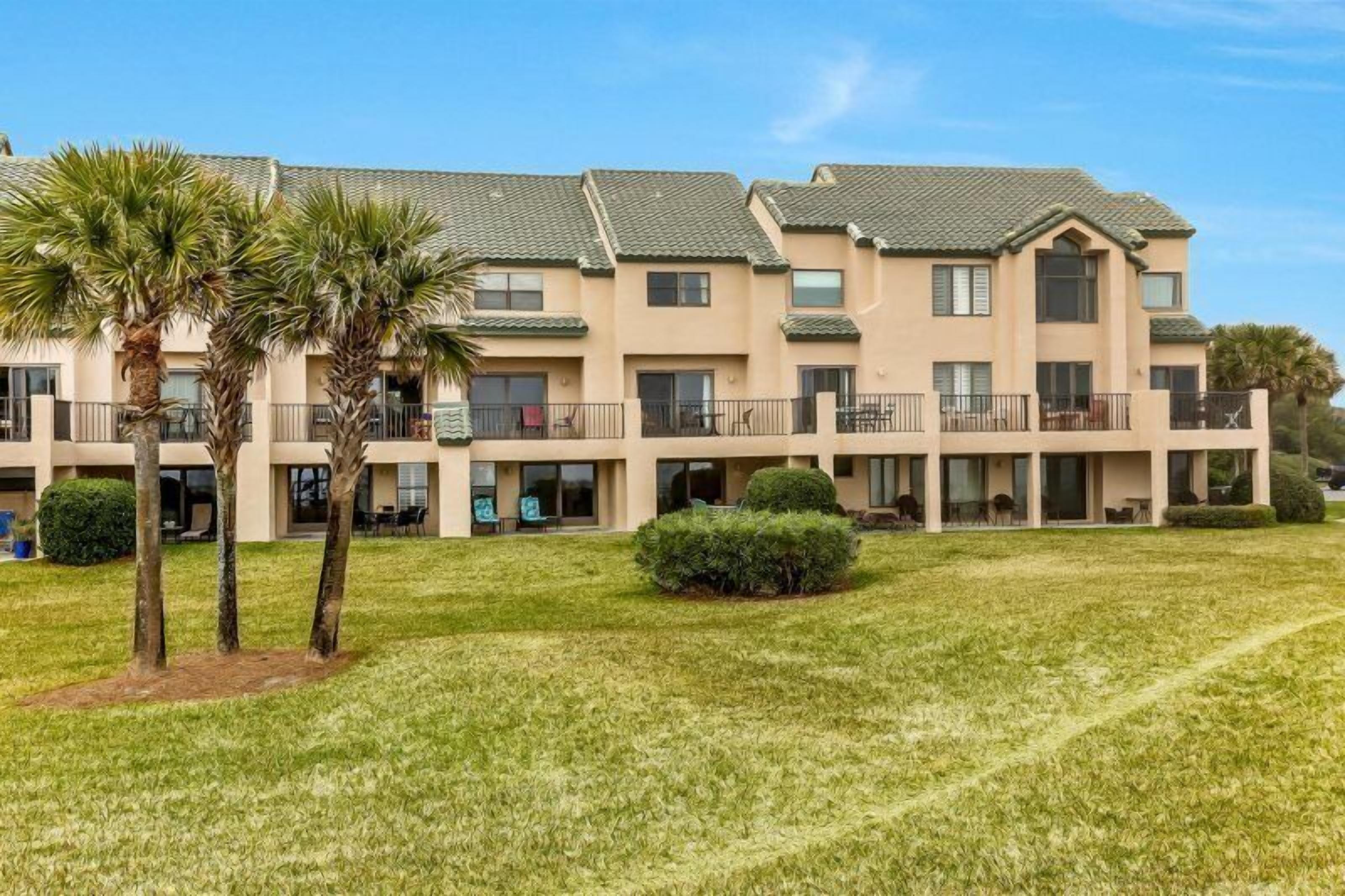 A multi-story hotel with a flat roof, balconies, and a well-maintained lawn with palm trees.