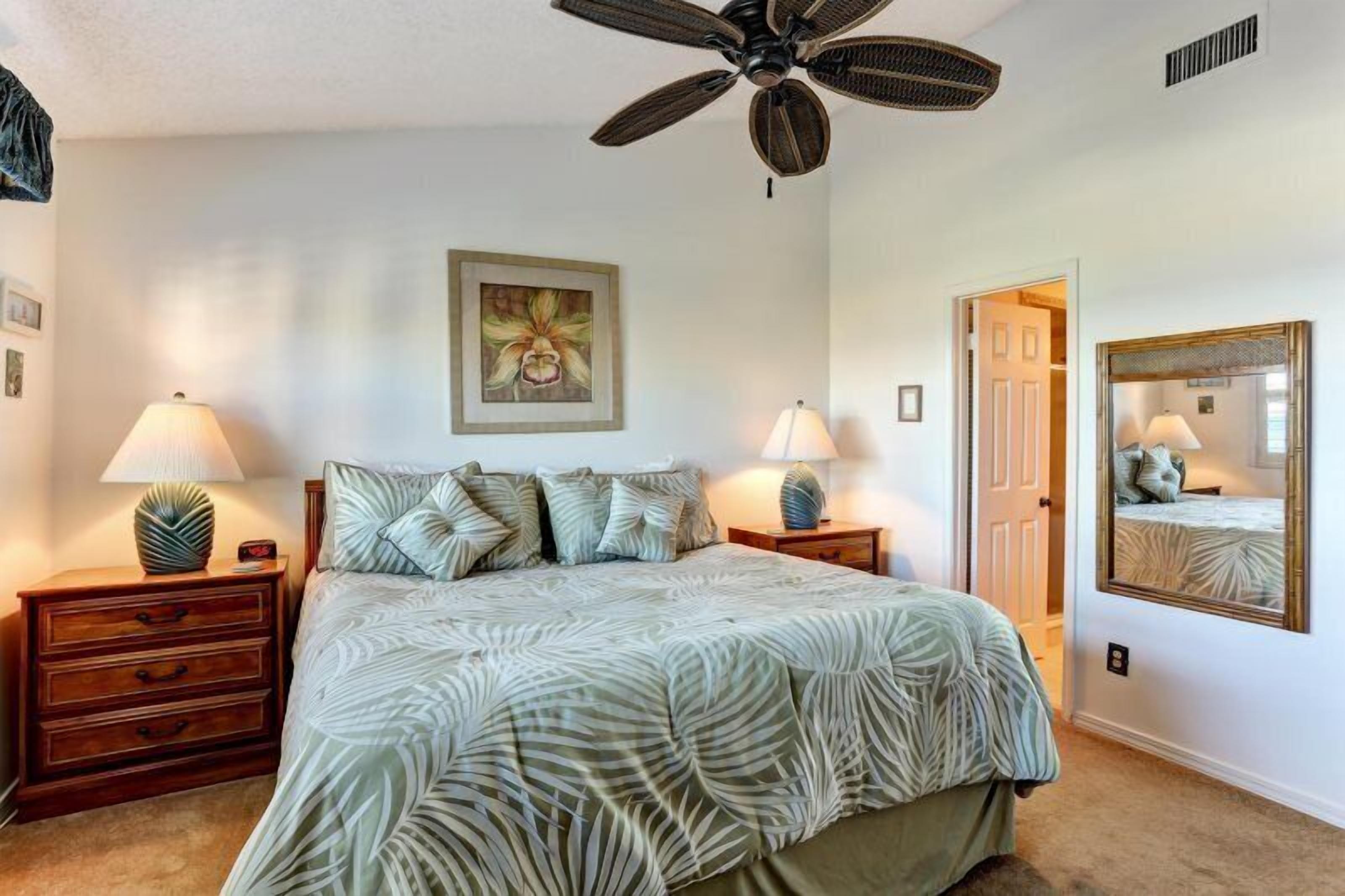 Une chambre à coucher comprenant un lit, deux tables de chevet, un ventilateur de plafond, un miroir fixé au mur et une porte donnant sur une autre pièce.