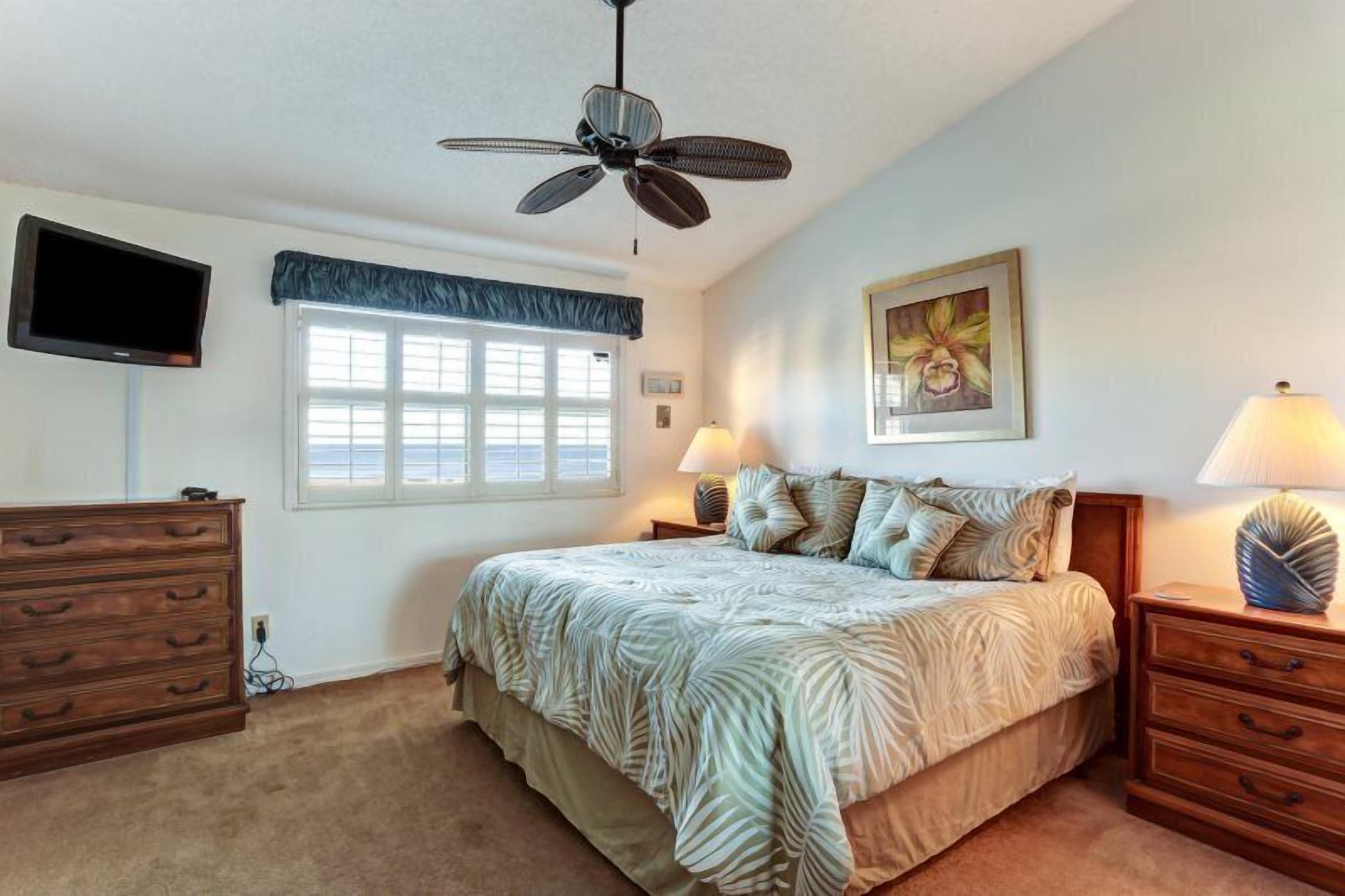 A bedroom with a bed, a ceiling fan, a TV mounted on the wall, a window with blinds, a dresser, and a lamp on the nightstand.