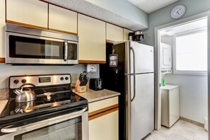 Kitchen features a stove, refrigerator, microwave, and kettle, with a door leading to another room.