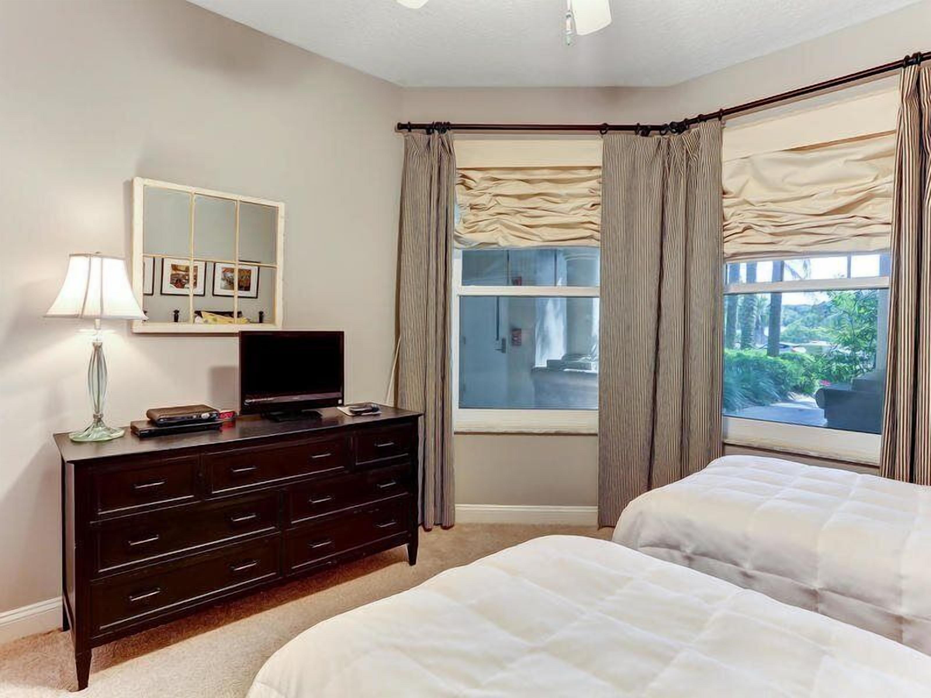A bedroom with two beds, a dresser, a TV, a mirror, and a window with curtains.
