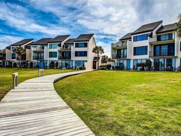 A modern multi-story residential building with a wooden deck leading to a grassy area.