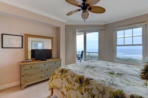 A bedroom with a bed, a TV on a dresser, a ceiling fan, and a balcony with a view of the ocean.
