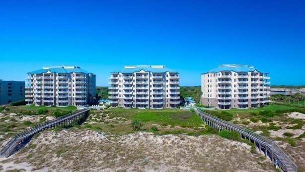 Three multi-story buildings with balconies, connected by a bridge, in a coastal area.