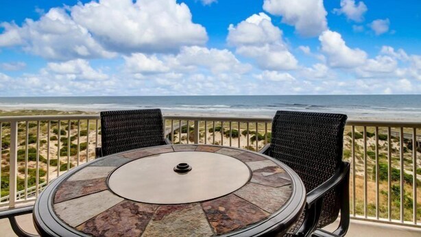 Mesa redonda con tapa de piedra y dos sillas de mimbre, con vistas a una playa y cielo despejado con nubes.