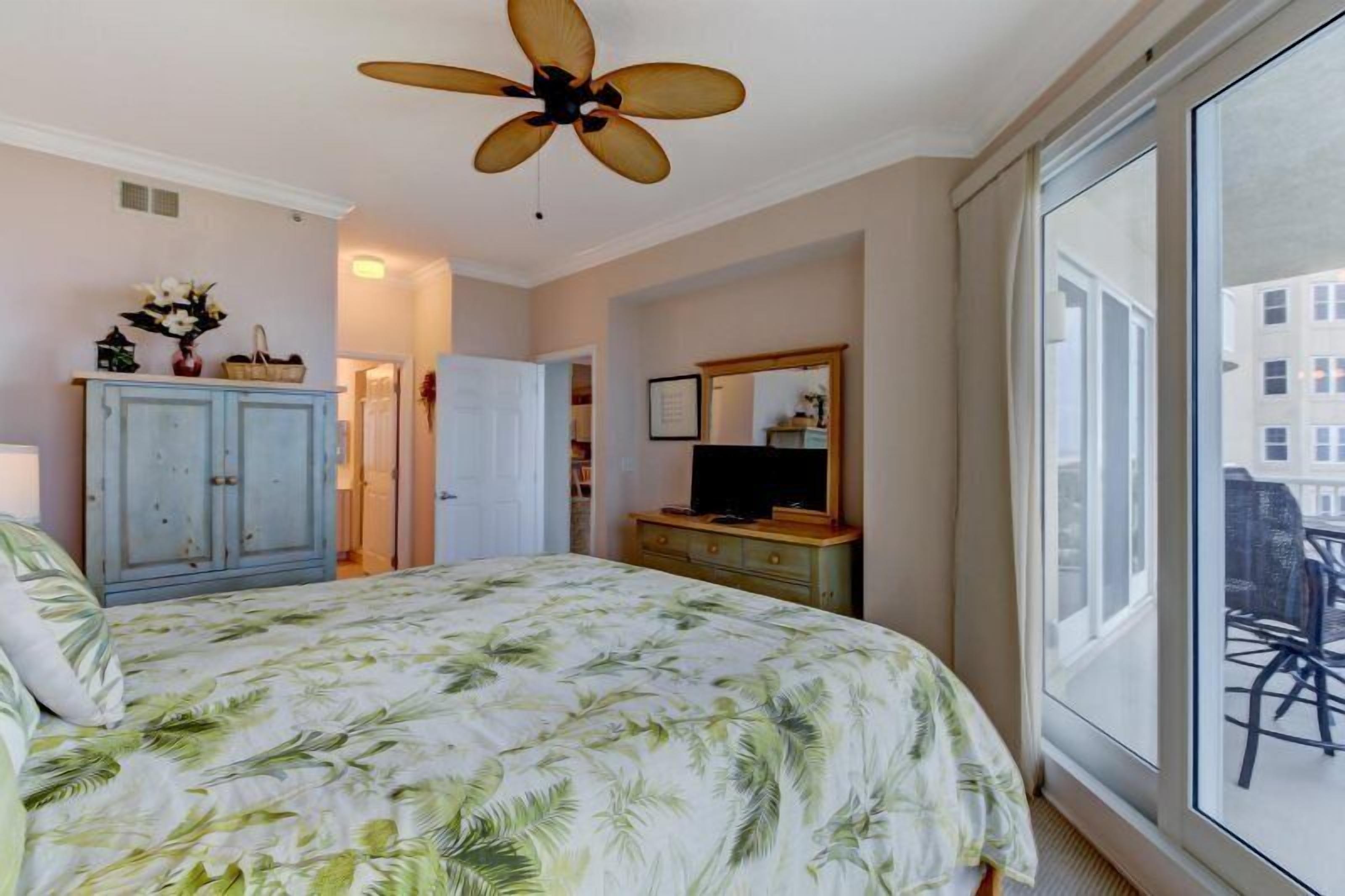 A bedroom with a bed, a ceiling fan, a dresser, a television, and a large window.