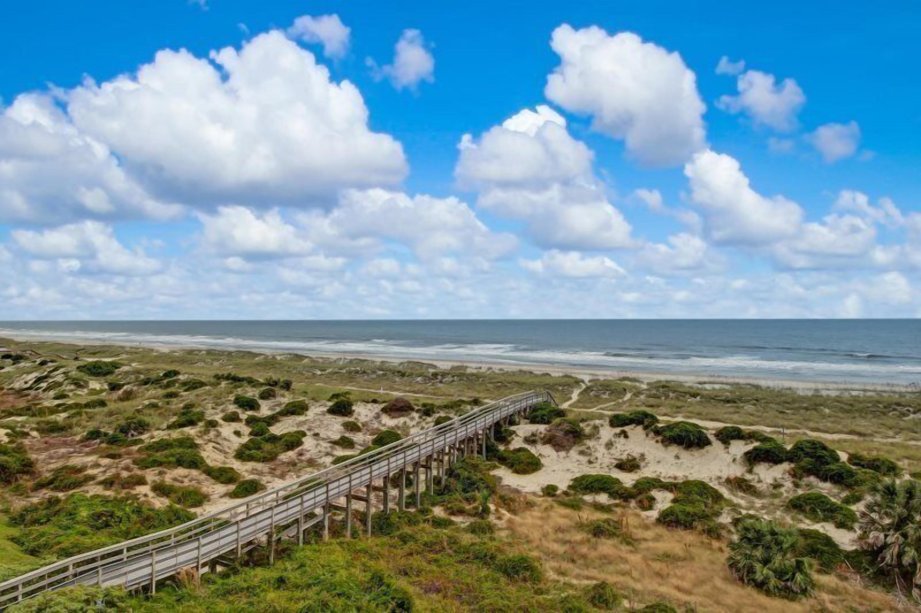 Eine Küstenlandschaft mit einem hölzernen Promenadenweg, der zum Strand führt, sandigen Dünen und einem klaren, blauen Himmel mit Wolken.