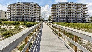 Un paseo marítimo de madera que conduce a un complejo de apartamentos frente a la playa con varios balcones.