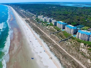 An aerial view of a coastal area with a beach, a road, and several high-rise buildings with blue roofs.