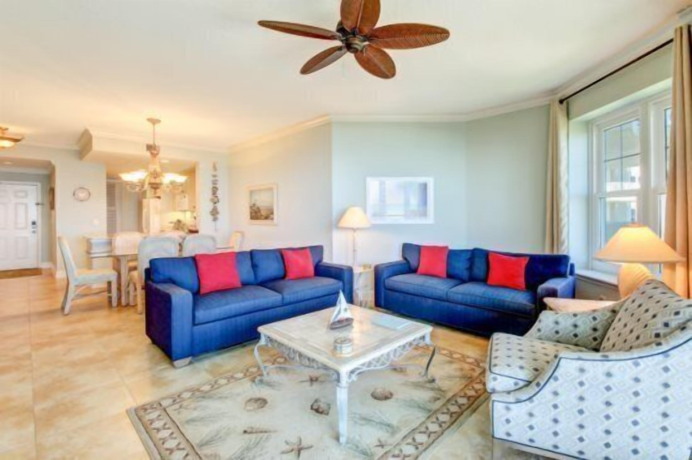 Un salon avec deux canapés bleus, un fauteuil à motifs, une table basse et un ventilateur de plafond.