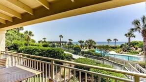 A balcony with a wooden table and chairs, overlooking a landscaped area with a pool and palm trees. - 1340-shipwatch (Fernandina Beach)