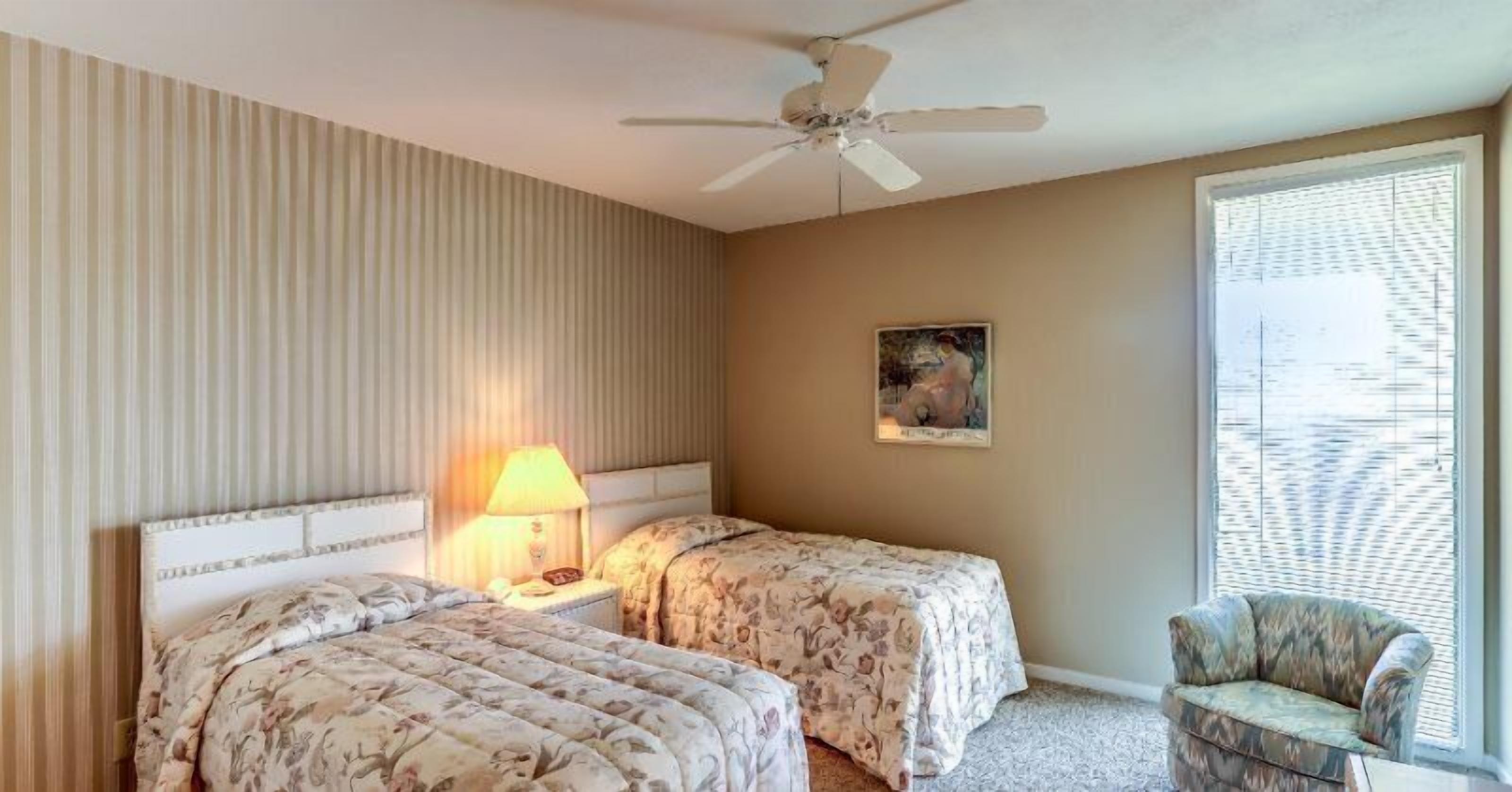 Ein Hotelzimmer mit zwei Betten, einem Deckenventilator, einer Lampe, einem Gemälde und einem Fenster mit Jalousien.
