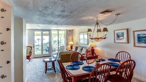 A dining area with a table set for four, surrounded by wooden chairs. There is a chandelier above the table, and the room has a view through large glass doors to an outdoor area. - 1340-shipwatch (Fernandina Beach)