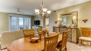 A dining area with a round wooden table, chairs, a chandelier, and a view of the kitchen in the background. - 42-ocean Place (Fernandina Beach)