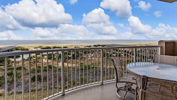 A balcony with a metal railing, a round table, and chairs, overlooking a beach with dunes and a clear sky. - 42-ocean Place (Fernandina Beach)