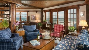 Un intérieur chaleureux avec un plafond en bois, de grandes fenêtres offrant une vue sur l’océan et divers types de sièges, notamment un canapé bleu à carreaux, un fauteuil rouge à motifs écossais et un divan à motifs floraux.