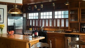 Une cuisine avec des meubles en bois, un îlot central agrémenté de verres à vin et une grande fen être avec des volets.