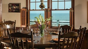 Une table à manger dressée pour quatre personnes, avec des chaises en bois, un vase de fleurs et une vue sur l’océan par de grandes fenêtres.