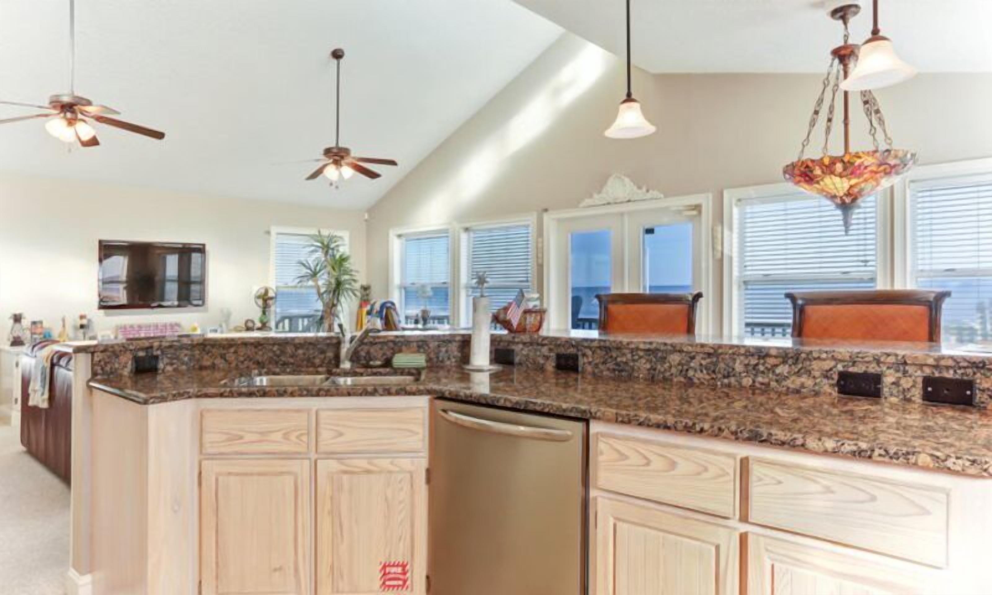 A spacious kitchen with granite countertops, wooden cabinets, and a central island. There are ceiling fans, pendant lights, and a large window providing natural light.