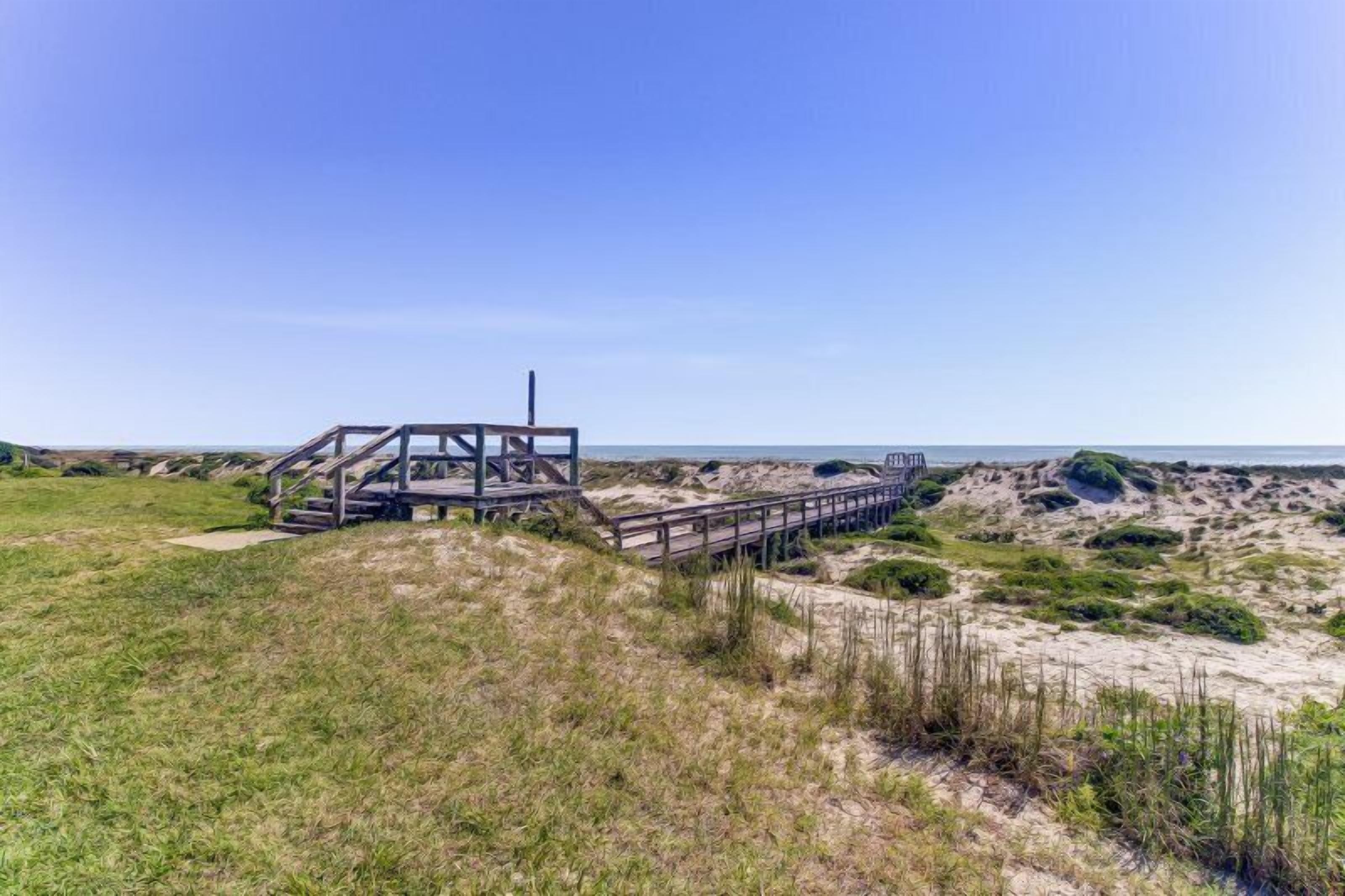 A wooden boardwalk leading to a simple, open structure with a staircase, surrounded by sandy dunes and sparse vegetation.
