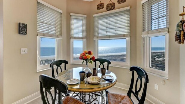 A dining area with a table set for four, four chairs, a vase with flowers, and a view of the ocean through large windows. - 25-ocean Place (Fernandina Beach)