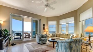 A spacious living room with a large sofa, two armchairs, a coffee table, and a ceiling fan. There is a sliding glass door leading to a balcony with a view of the ocean. - 25-ocean Place (Fernandina Beach)
