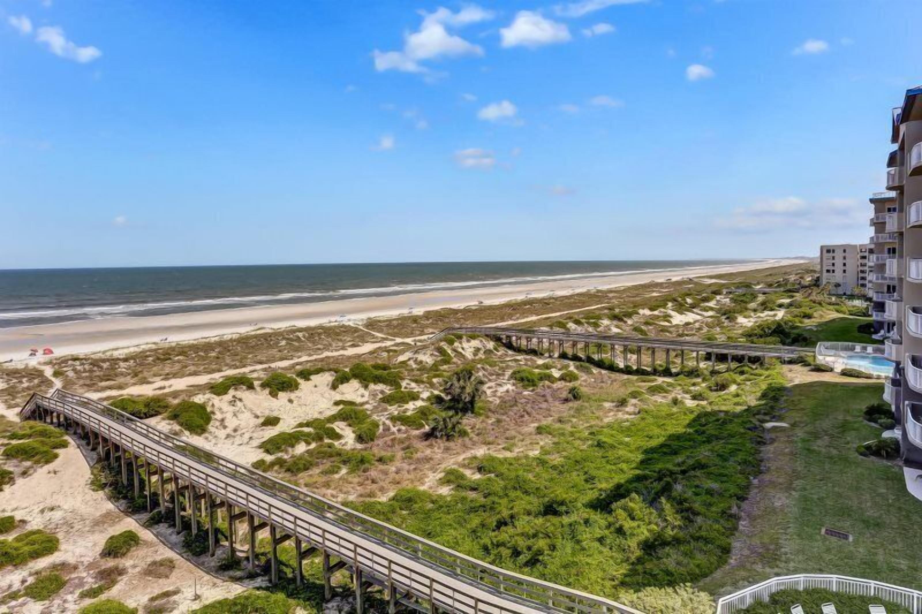 Blick auf einen Strand mit einer Holzsteg, Sanddünen und einem Gebäude mit Balkonen mit Meerblick.