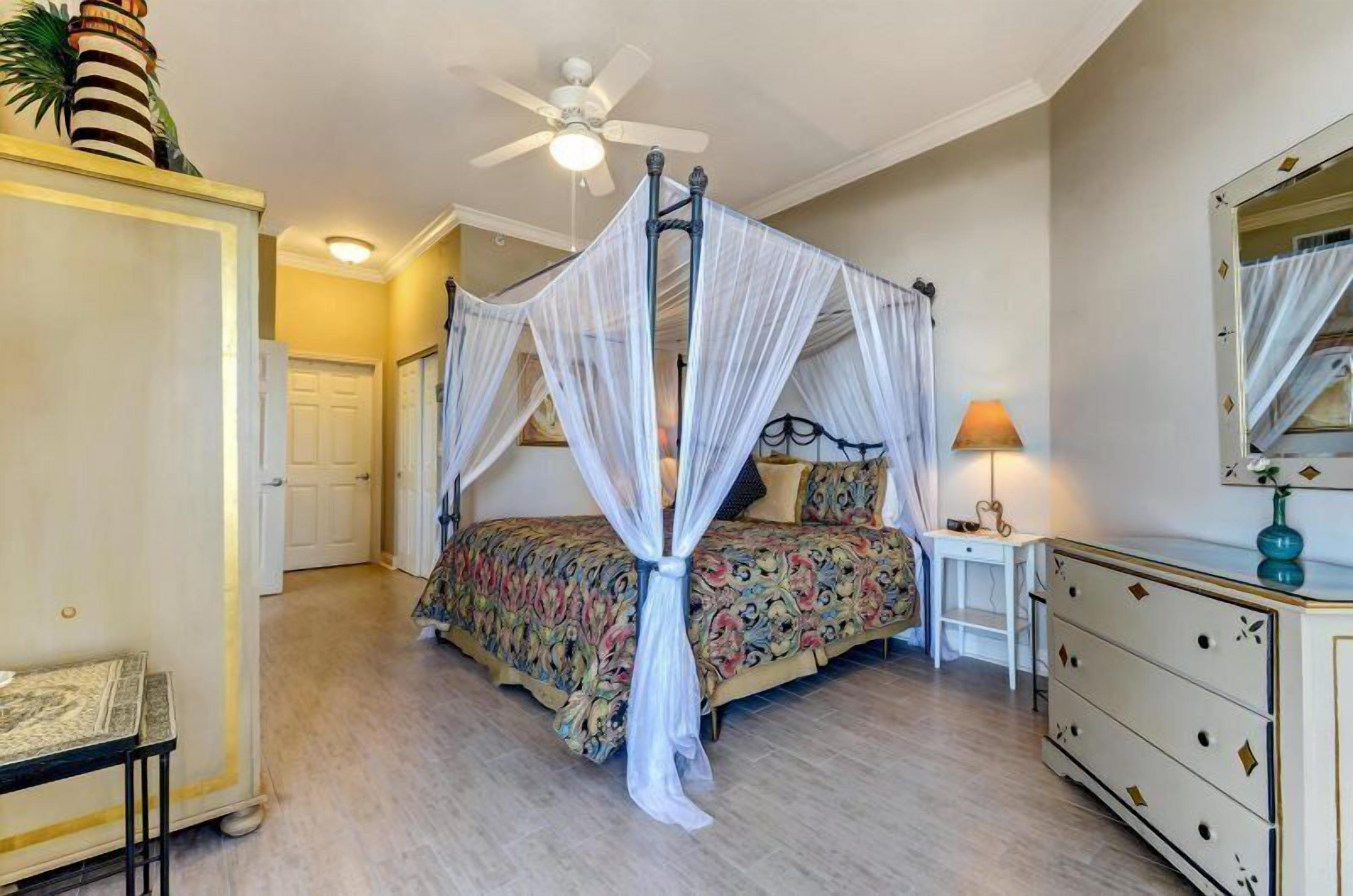 A bedroom with a four-poster canopy bed, a bedside table with a lamp, and a dresser with drawers.
