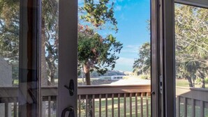 A view from a balcony with a wooden railing, a door with a handle, and a clear sky with trees and a distant landscape. - 2098-beachwood (Fernandina Beach)