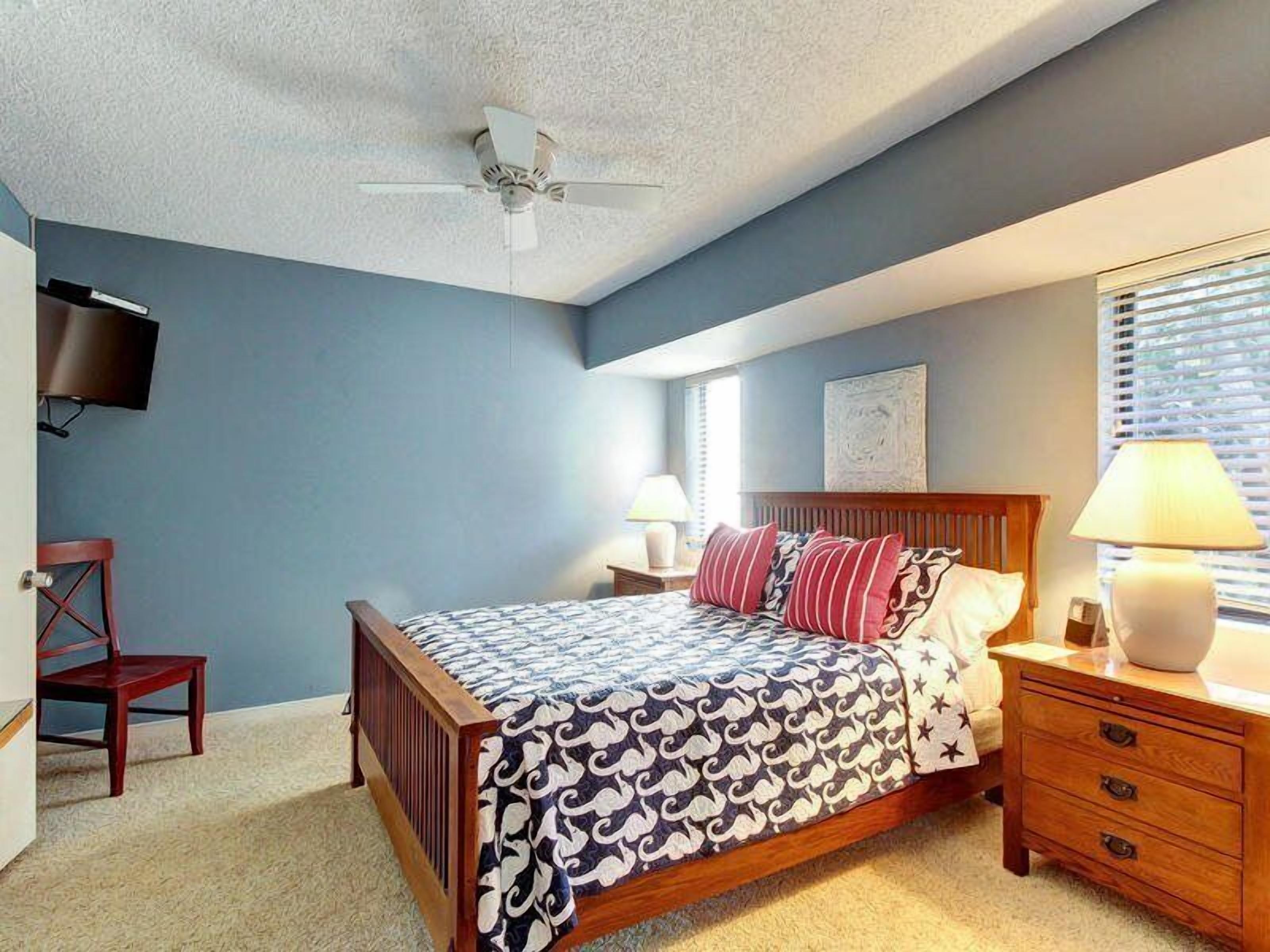 A bedroom with a bed, bedside tables, a chair, a ceiling fan, and a TV mounted on the wall.
