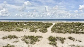 Ein Strand mit einem sandigen Pfad, der zum Wasser führt, unter einem teils bewölkten Himmel.