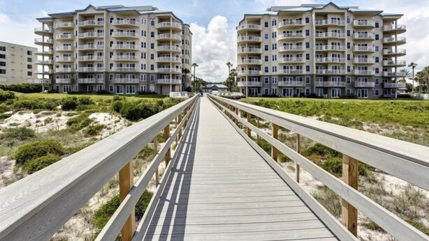 Ein hölzerner Steg führt zu einem Apartmentkomplex direkt am Strand, mit mehreren Balkonen und blauem Himmel.
