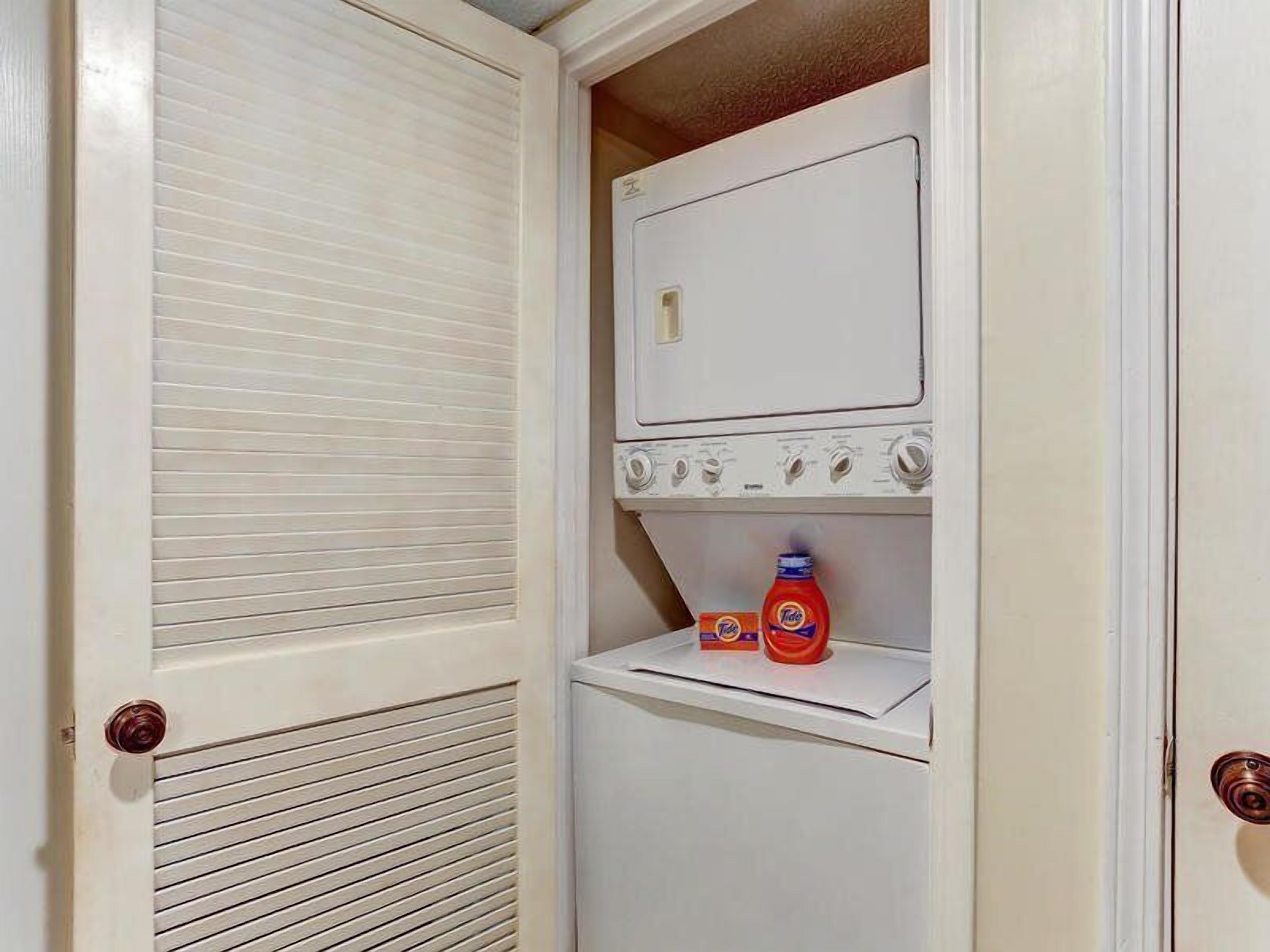 A laundry room with a stacked washer and dryer, a shelf with Tide detergent, and a door with a louvered design.