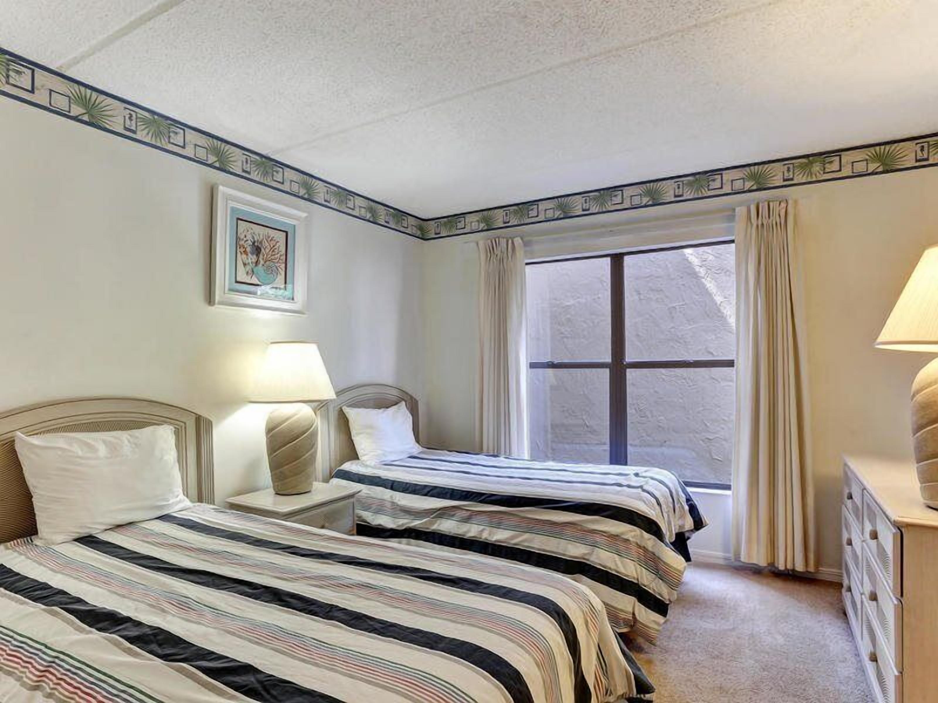 A hotel room with two beds, a window with curtains, a bedside table with a lamp, and a wall-mounted picture.