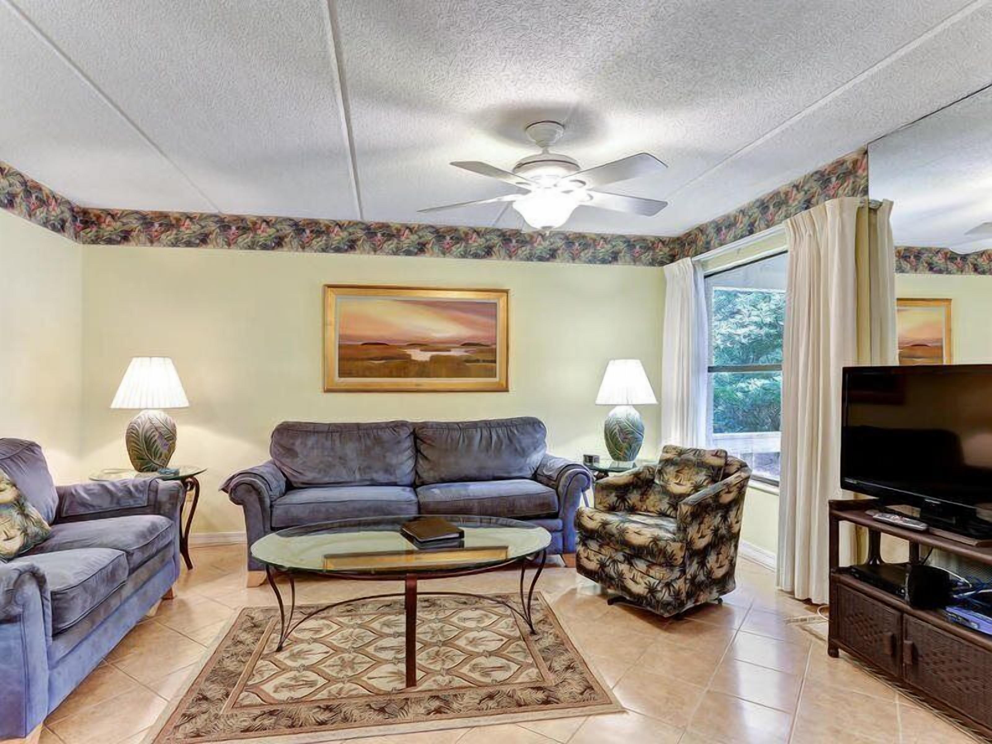 A living room with a ceiling fan, two sofas, a coffee table, a patterned armchair, and a television.