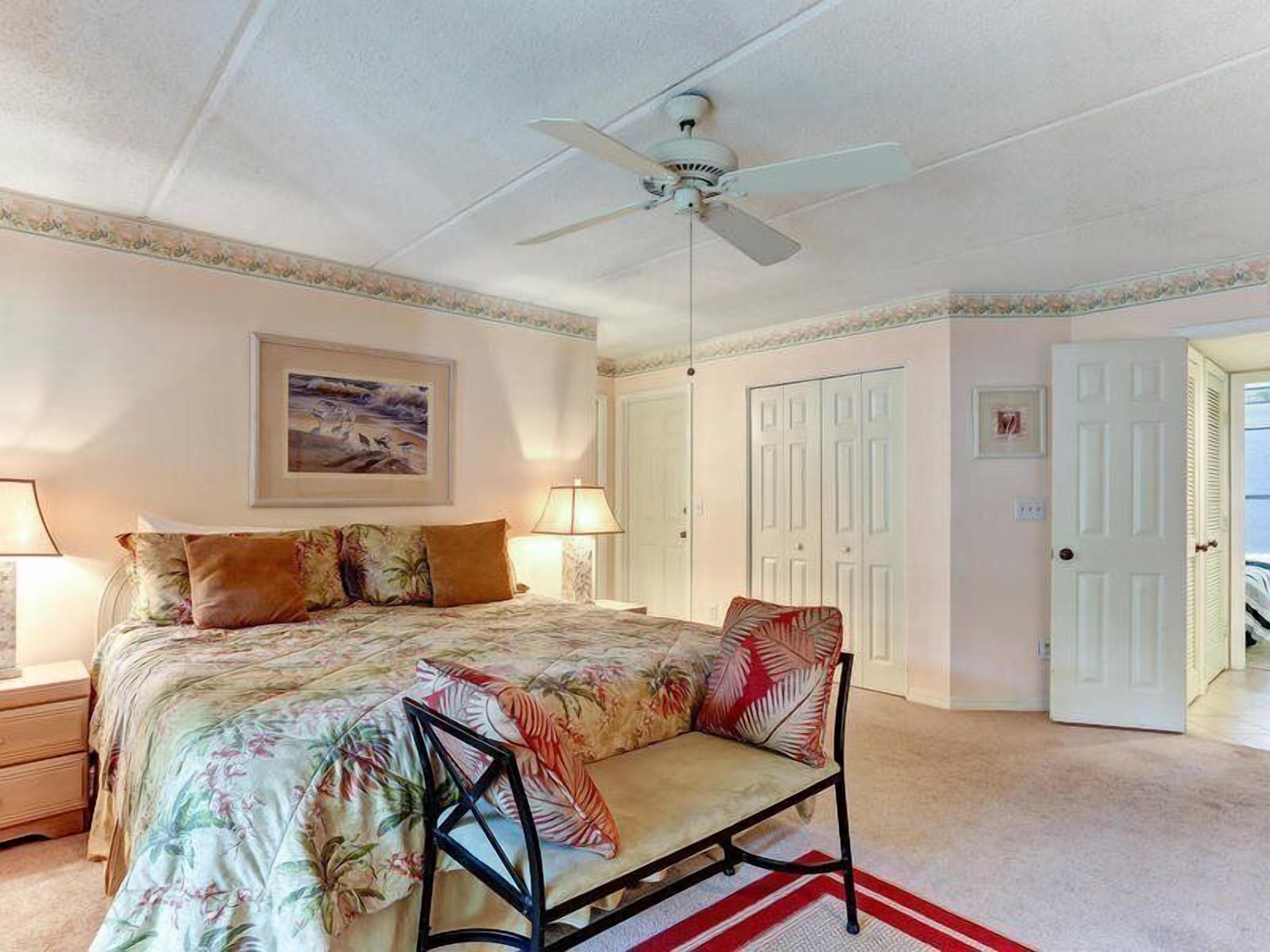 Une chambre à coucher comprenant un lit, une chaise, un ventilateur de plafond, une table de chevet, une lampe et une porte.