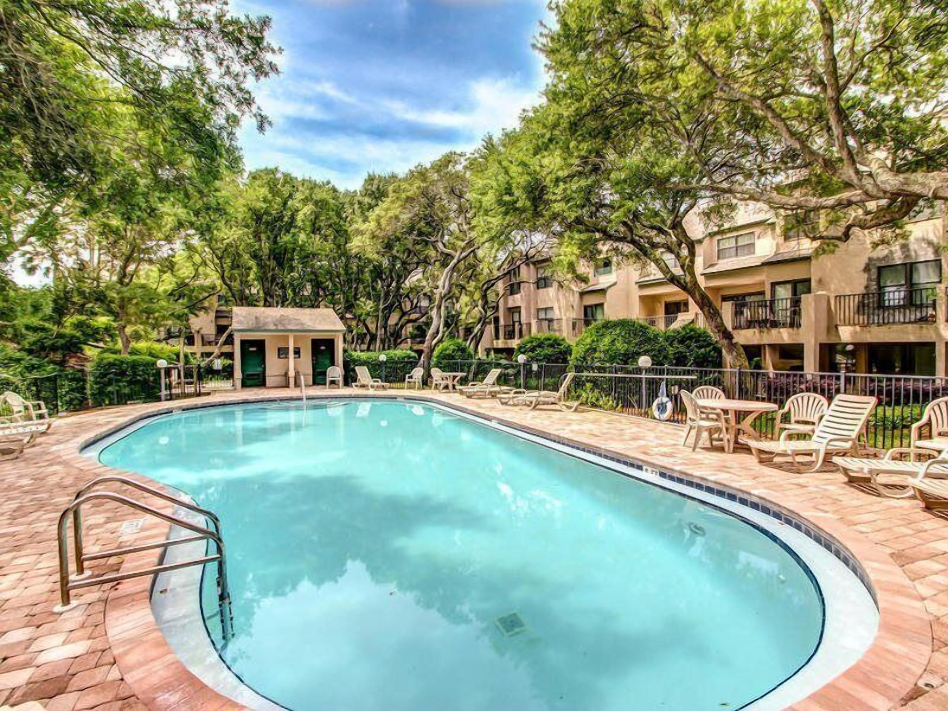 Une piscine avec des chaises longues et un petit bâtiment, le tout entouré d’arbres, ainsi qu’un autre bâtiment doté de balcons.