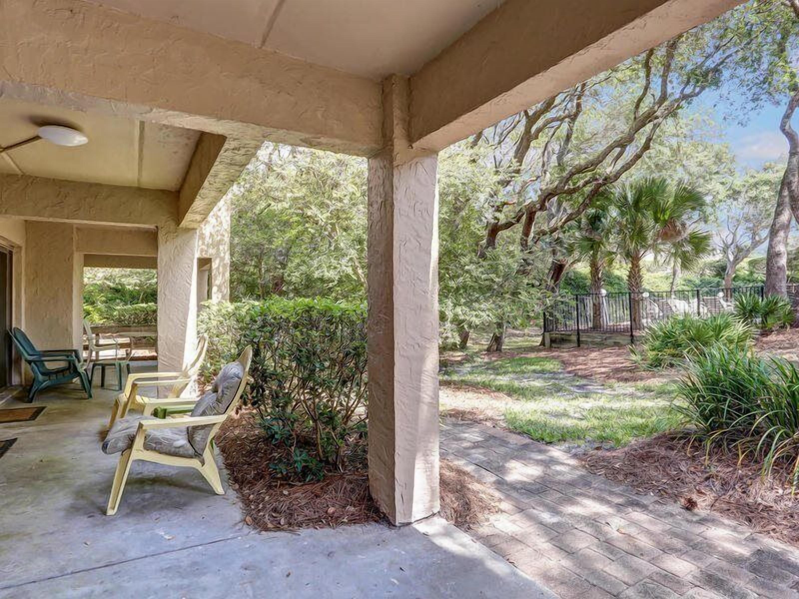 A covered patio area with outdoor furniture, a view of a landscaped garden, and a fence in the background.