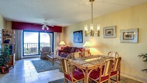 A living room with a dining table, chairs, a sofa, and a ceiling fan. There are two lamps on the table, and a window with curtains. - 1830-turtle-dunes (Fernandina Beach)