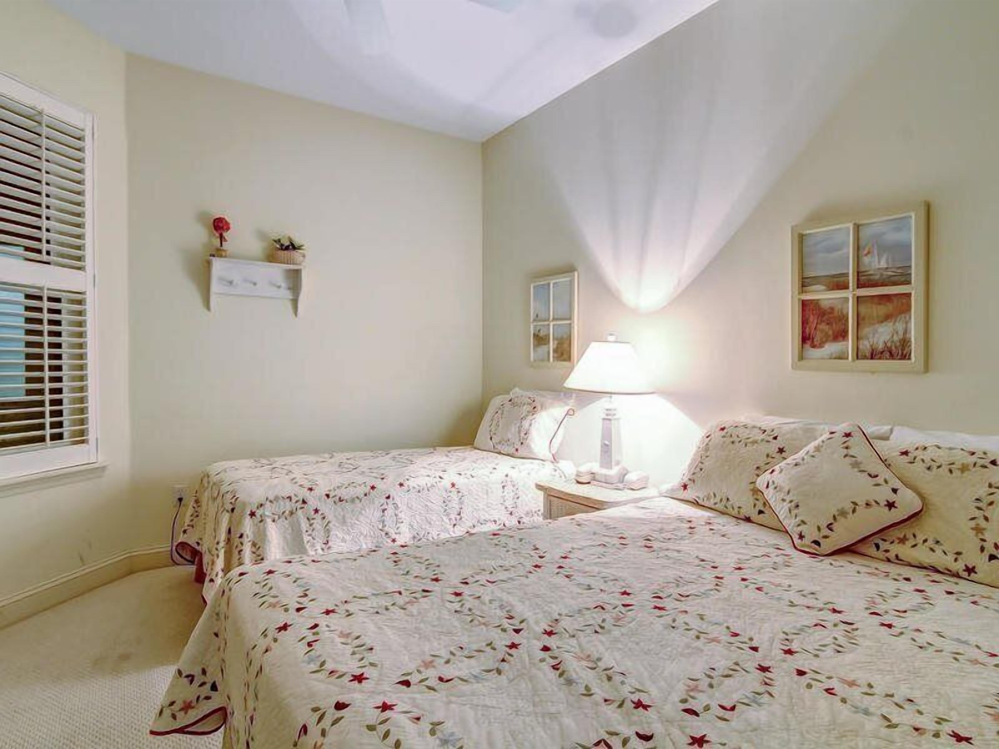 A bedroom with two beds, a nightstand, a lamp, a window with blinds, and a wall-mounted shelf with decorative items.
