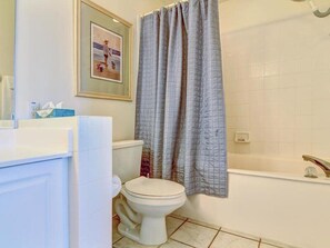 A bathroom with a toilet, a shower curtain, a framed picture on the wall, and a white sink with a mirror.