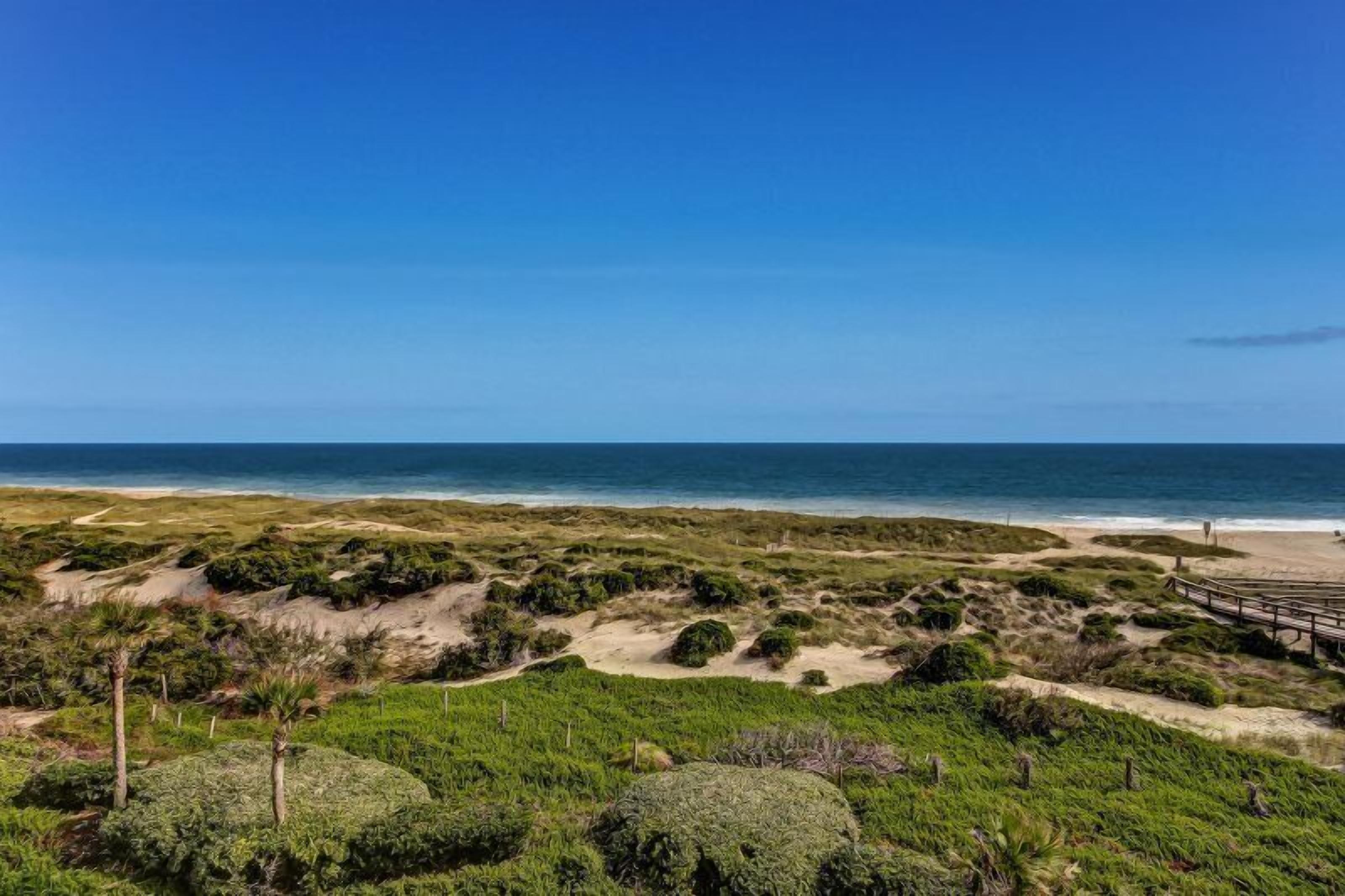 A coastal landscape with a sandy beach, green vegetation, and a wooden walkway leading to the shore.